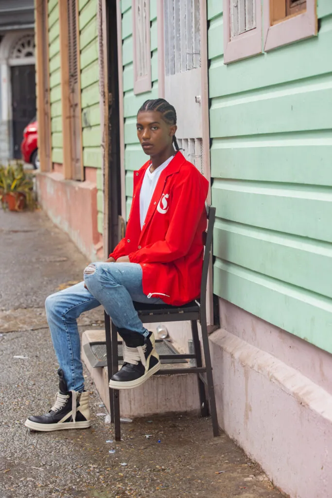 Male model in red jacket and blue jeans sitting on chair against pastel buildings