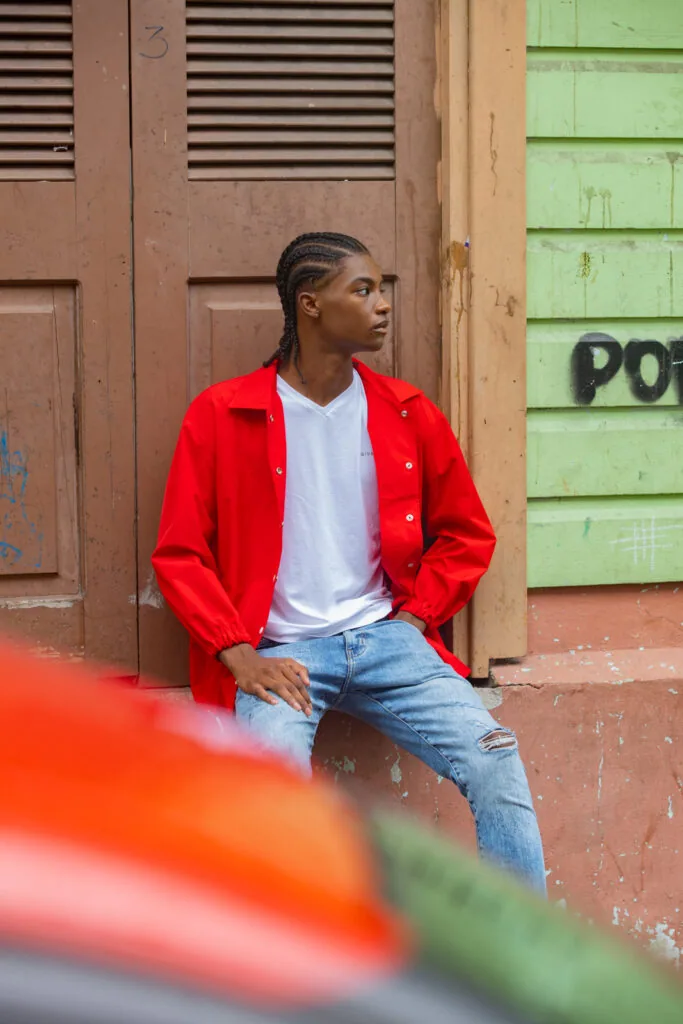 Male model in red jacket and white shirt seated against weathered brown and green doors in urban setting