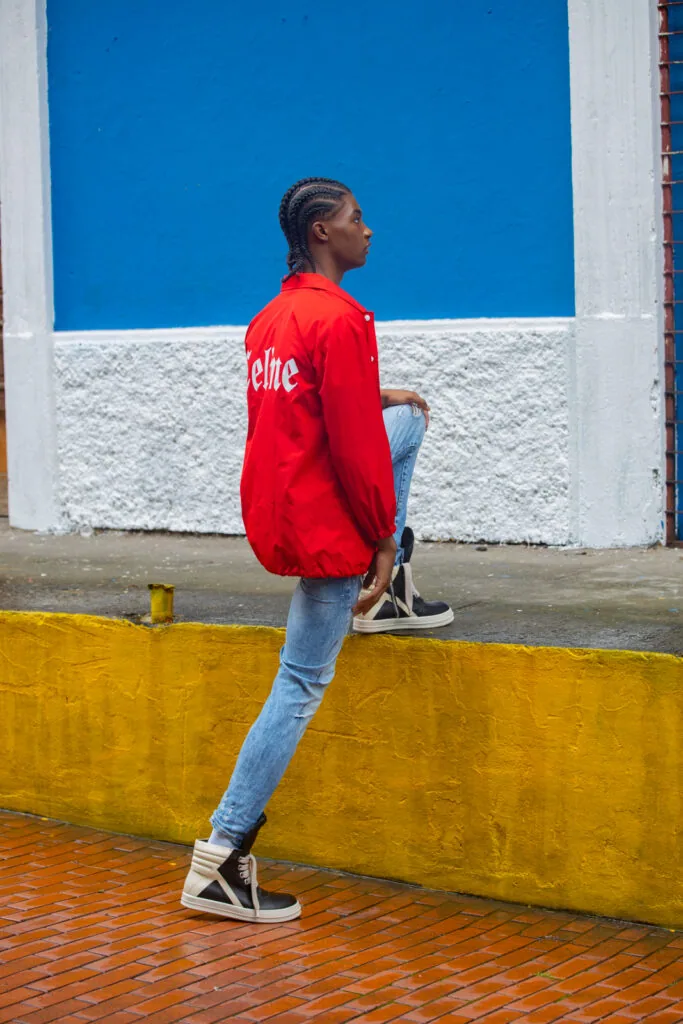 Man in red jacket and blue jeans posing against blue and white wall with yellow barrier