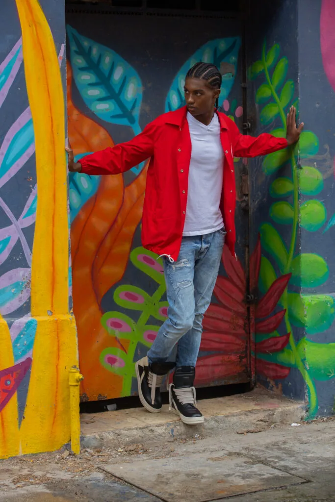 Male model in red jacket and white shirt posing with arms outstretched against colorful street art mural with tropical leaf designs.
