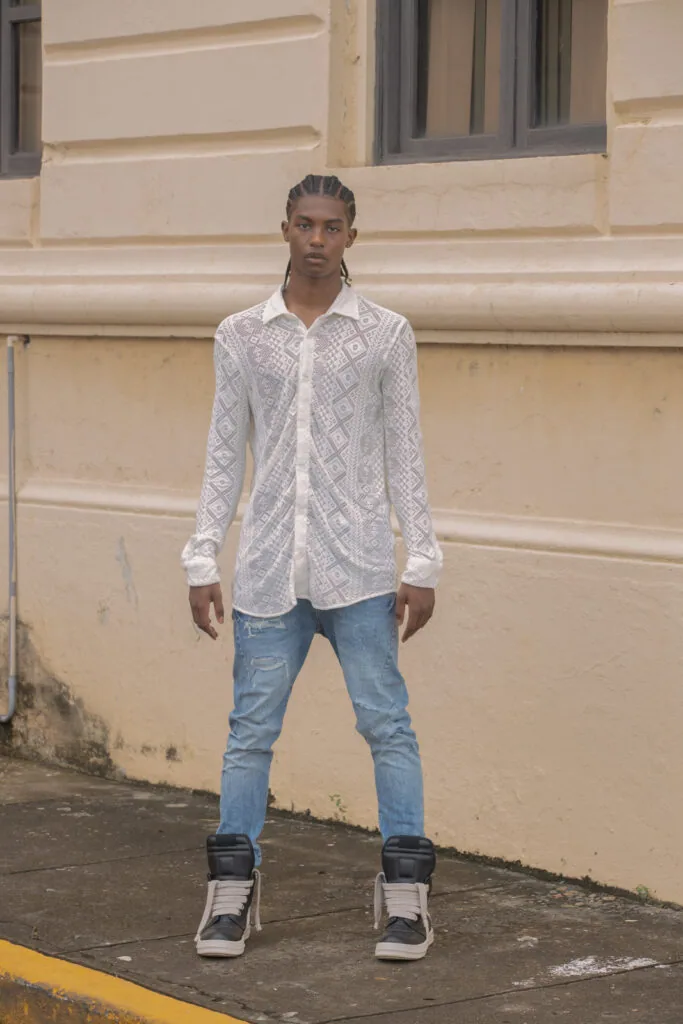 Male model wearing white geometric patterned shirt and light blue jeans with black and white high-top sneakers, standing against tan building facade