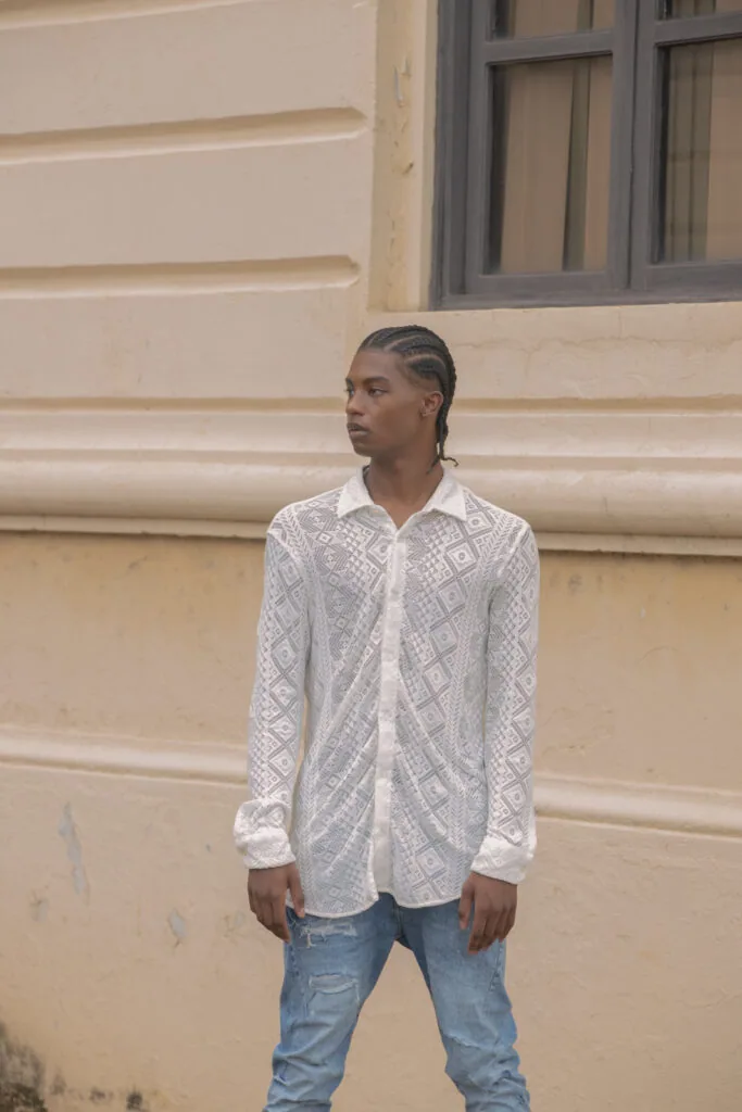 Male model wearing white patterned shirt and distressed blue jeans against beige building facade.