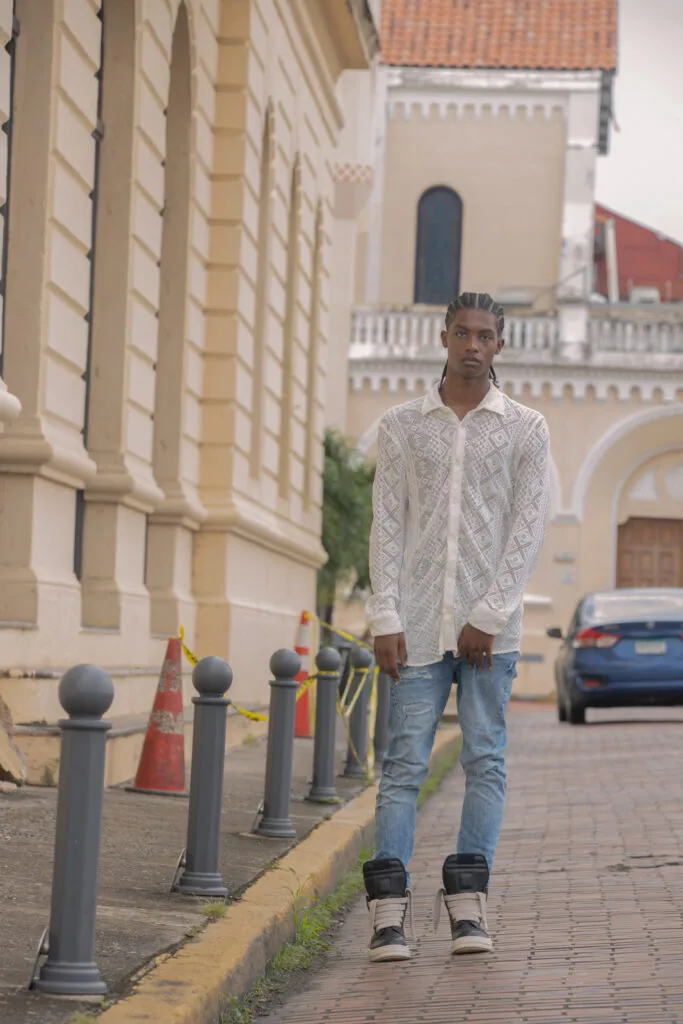 Male model in white patterned shirt and blue jeans standing on brick street with classical architecture backdrop