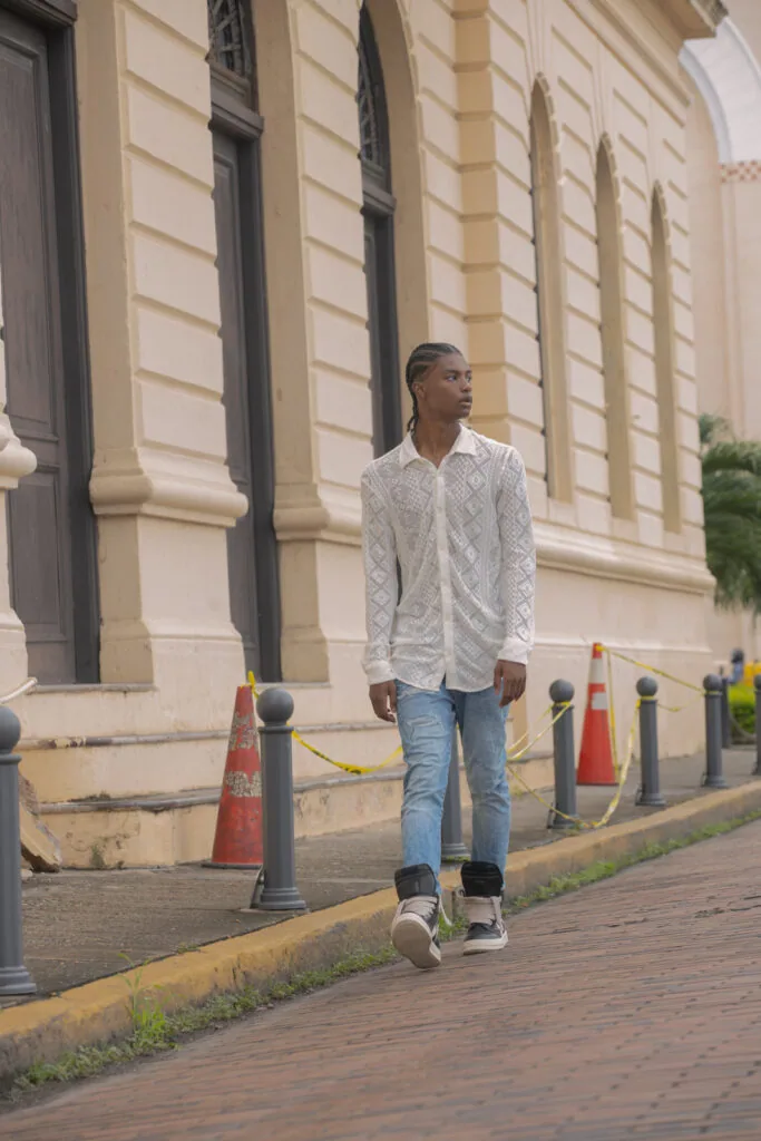 Man in white quilted shirt and blue jeans walking past classical stone building with columns and metal bollards