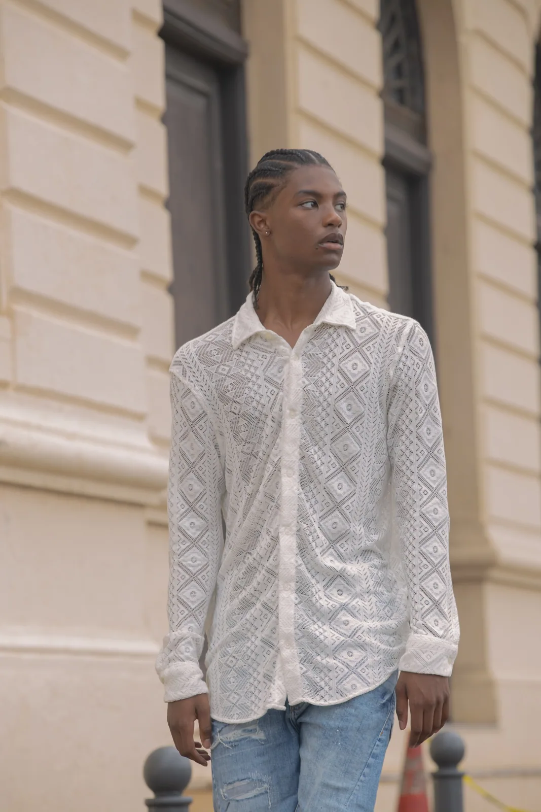 Male model wearing white geometric patterned shirt and distressed blue jeans against beige building facade with dark windows