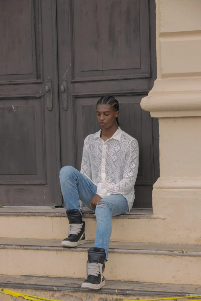Man sitting on steps wearing white patterned shirt, ripped blue jeans, and black sneakers against dark wooden doors
