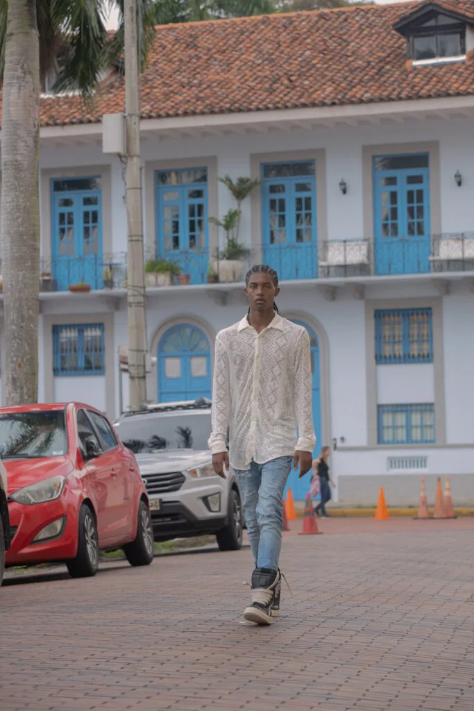 Male model in cream cable-knit shirt and jeans walking on brick plaza with blue colonial building and parked cars in background