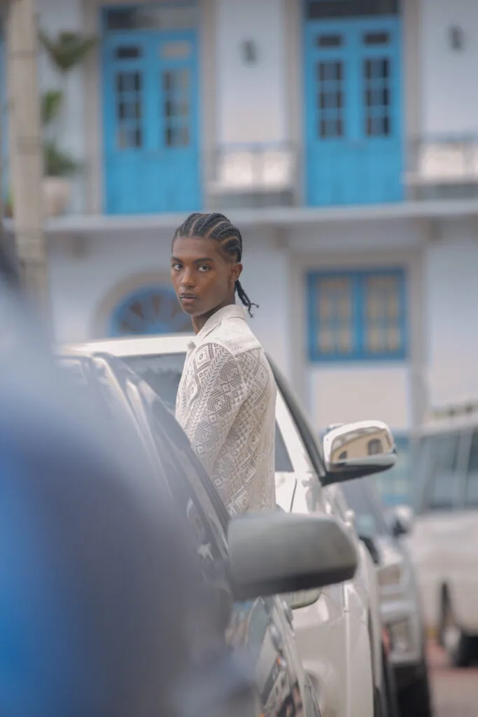 Male model in white knit sweater leaning against vehicle with blue-shuttered Mediterranean architecture backdrop
