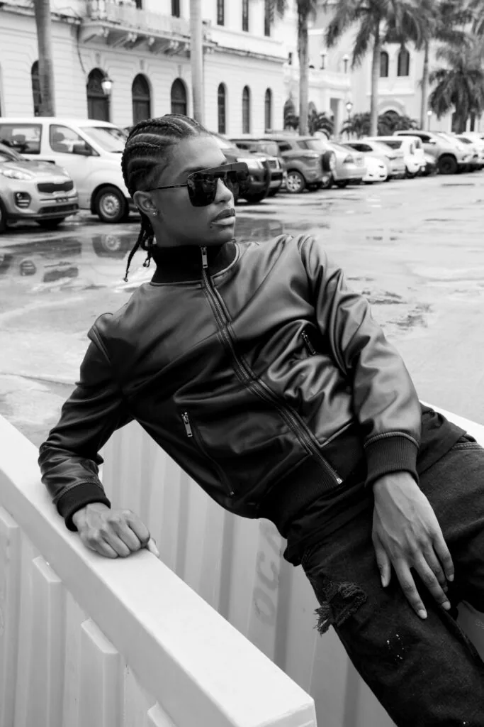 Male model in black leather jacket and sunglasses posing in parking lot with palm trees and white building