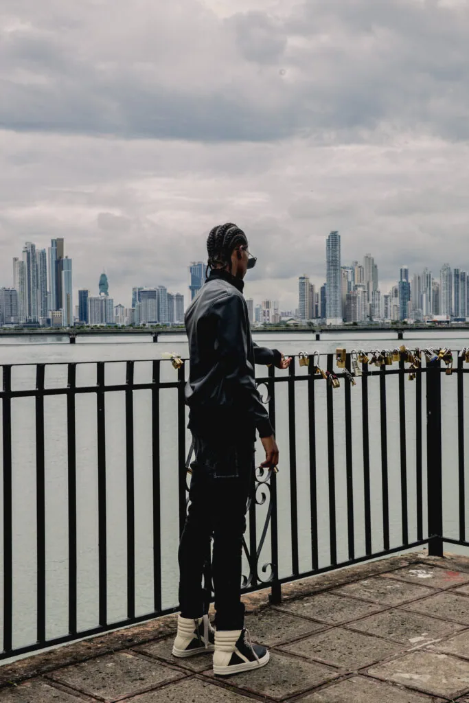 Male model in black jacket and white sneakers standing at waterfront railing overlooking city skyline across water.