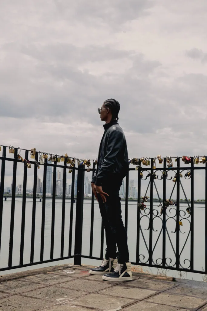 Male model in black jacket and pants standing on bridge viewing platform with metal railings and cloudy sky backdrop