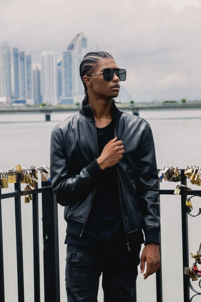Male model wearing black leather jacket and sunglasses standing at waterfront railing with city skyline behind