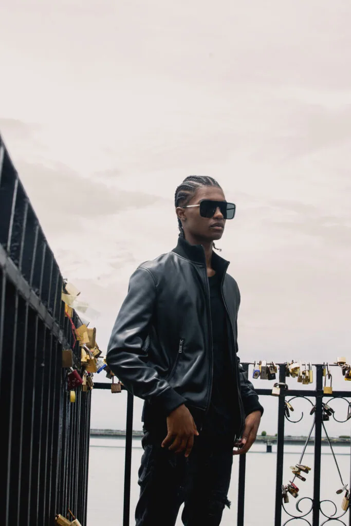 Male model in black leather jacket and sunglasses standing at metal railing overlooking water with overcast sky
