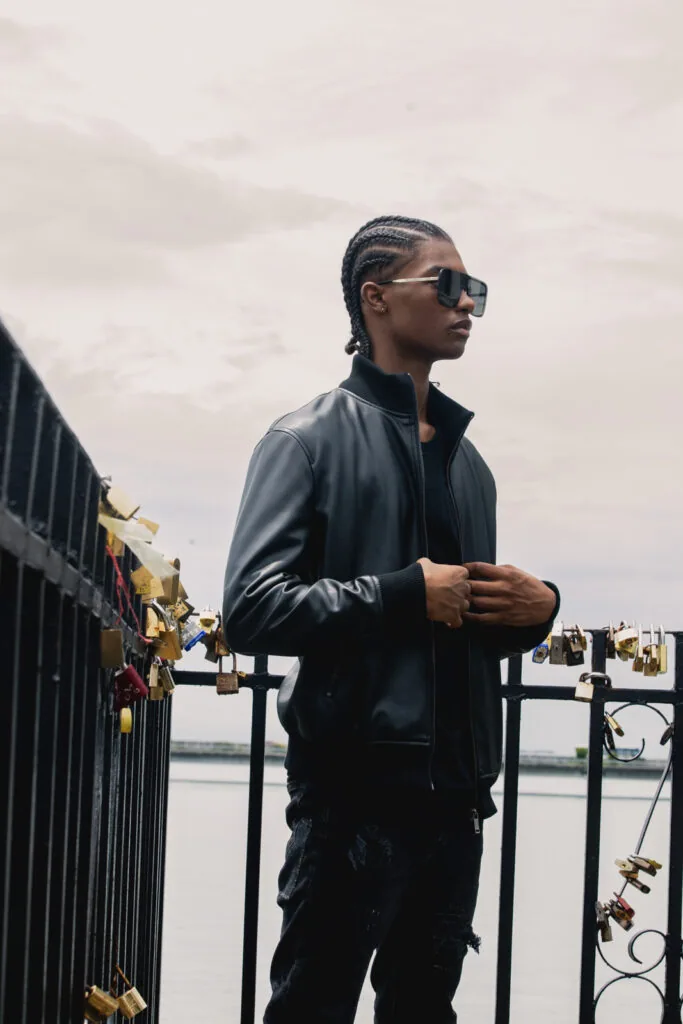 Male model wearing black leather jacket and sunglasses on waterfront bridge with grey sky backdrop