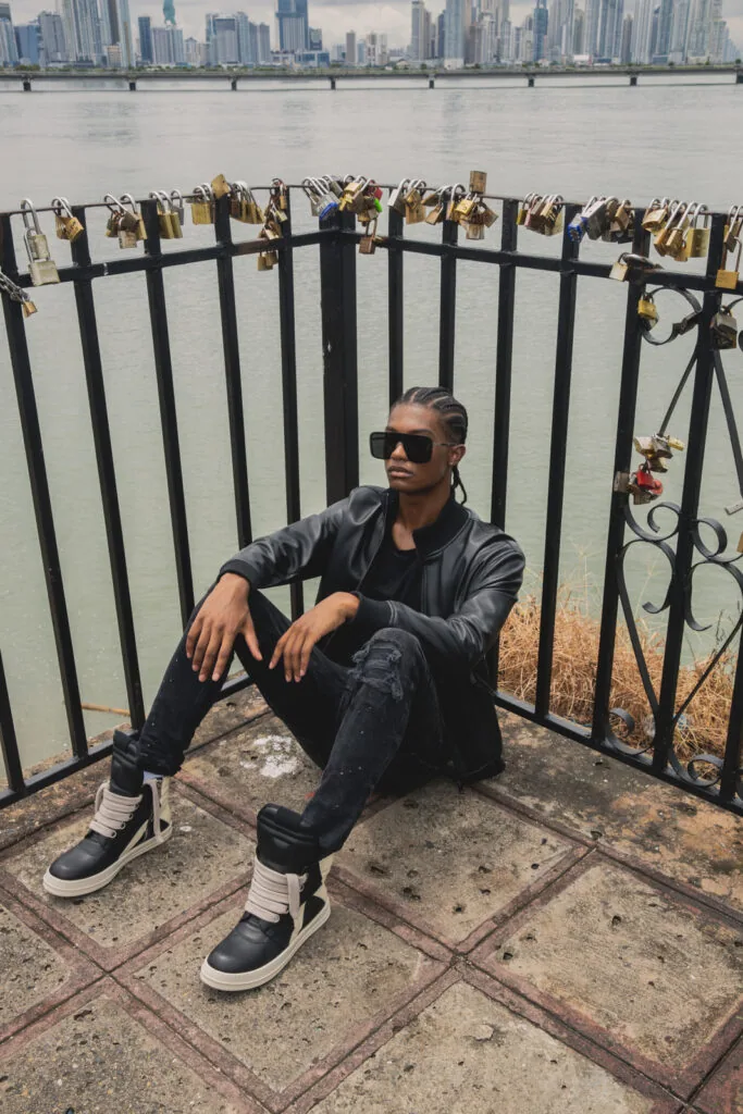 Man in black leather jacket and jeans squatting by railing with padlocks overlooking city skyline and water