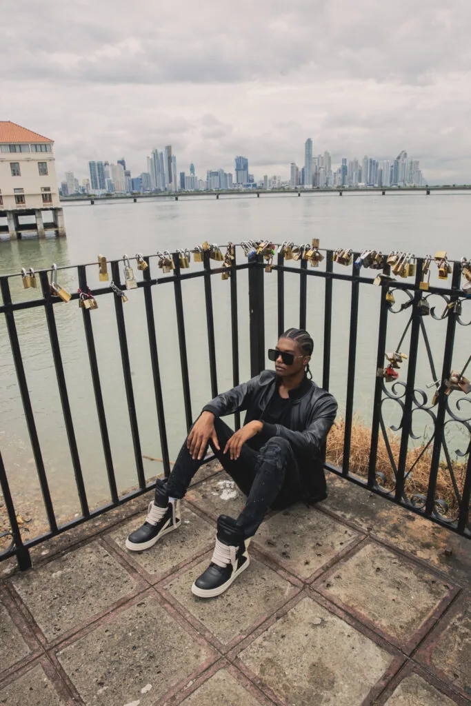 Man in black jacket and sunglasses sitting by waterfront railing with city skyline and love locks in background