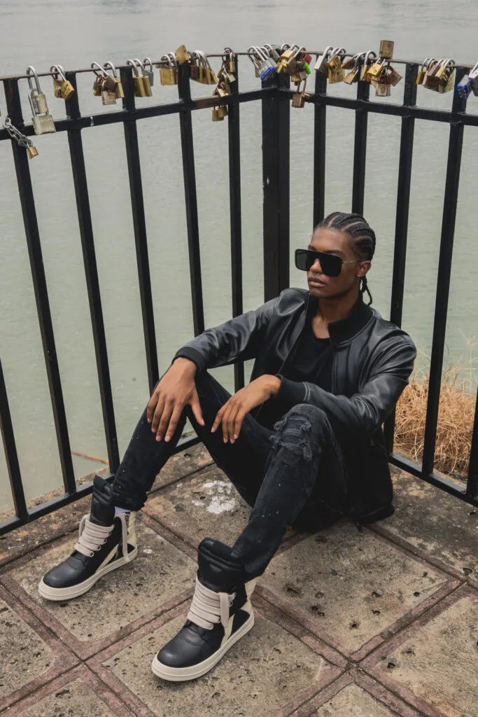 Male model in black jacket and distressed jeans with high-top sneakers posing against metal fence with padlocks