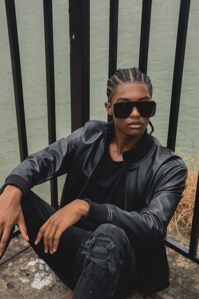 Man in black leather jacket and ripped jeans wearing dark sunglasses, sitting against vertical black fence with water background.