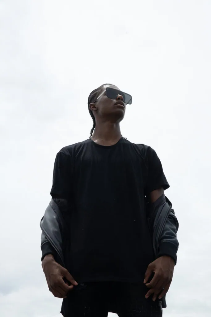 Male model wearing black t-shirt and gray jacket with sunglasses against cloudy sky