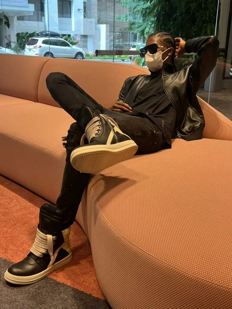 Man in black leather jacket and sneakers reclines on tan modern couch wearing dark sunglasses indoors.