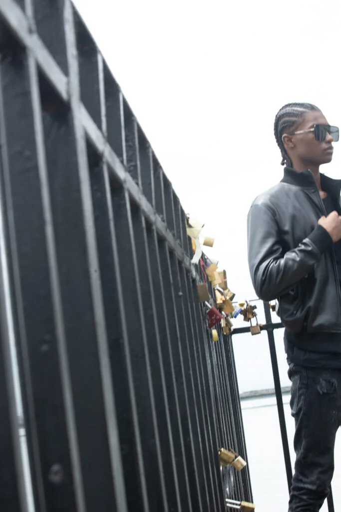 Male model in black leather jacket and sunglasses standing against industrial metal fence with sky background