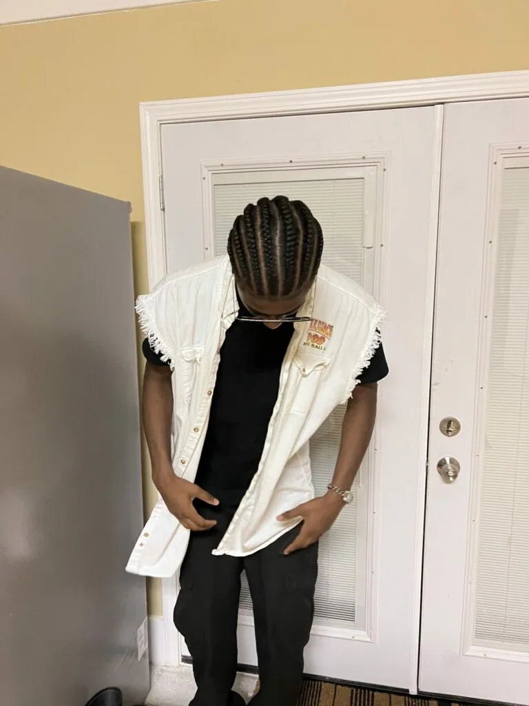Person wearing white sleeveless shirt over black t-shirt with cornrow braids, standing in front of white closet doors with yellow walls.