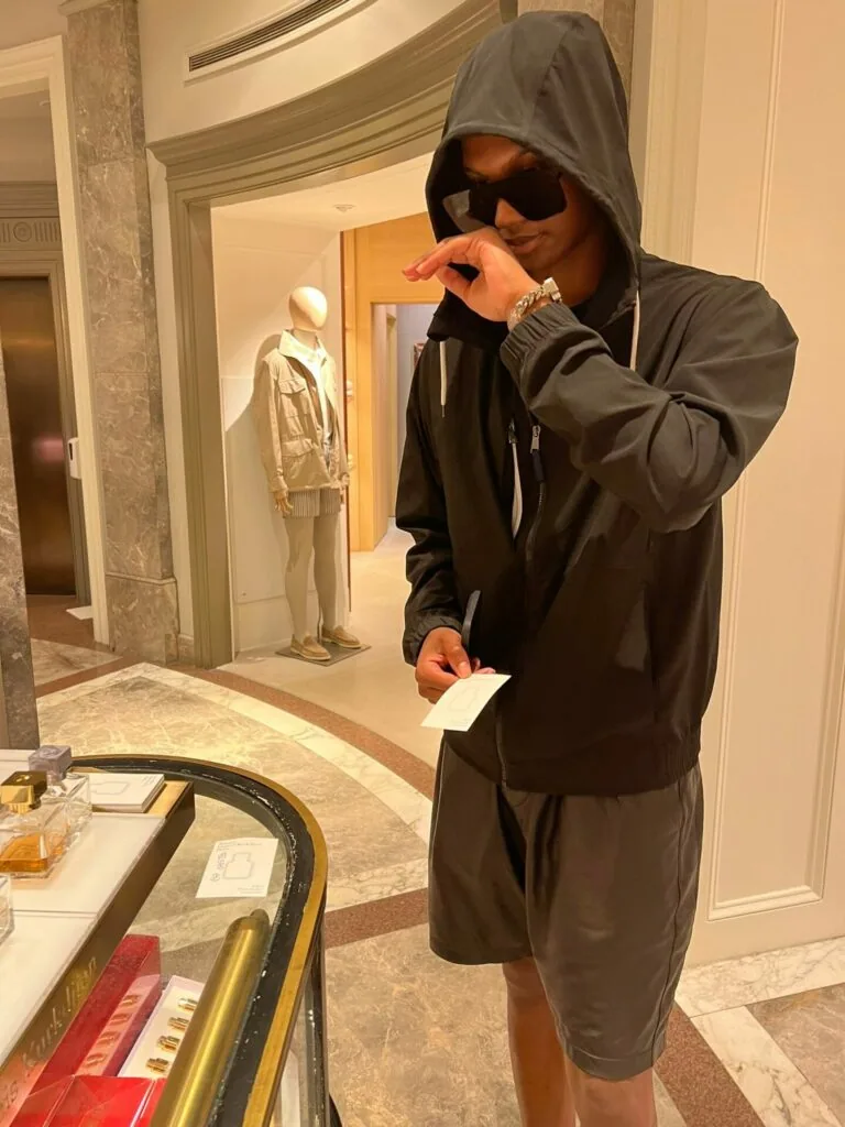 Man in black hoodie and sunglasses posing in gallery space with mannequin and artwork displays.