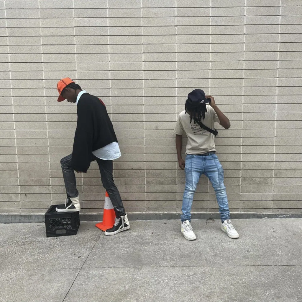 Two people pose against brick wall; person left wears orange cap, black jacket, grey pants; person right wears beige shirt, blue jeans, black cap.