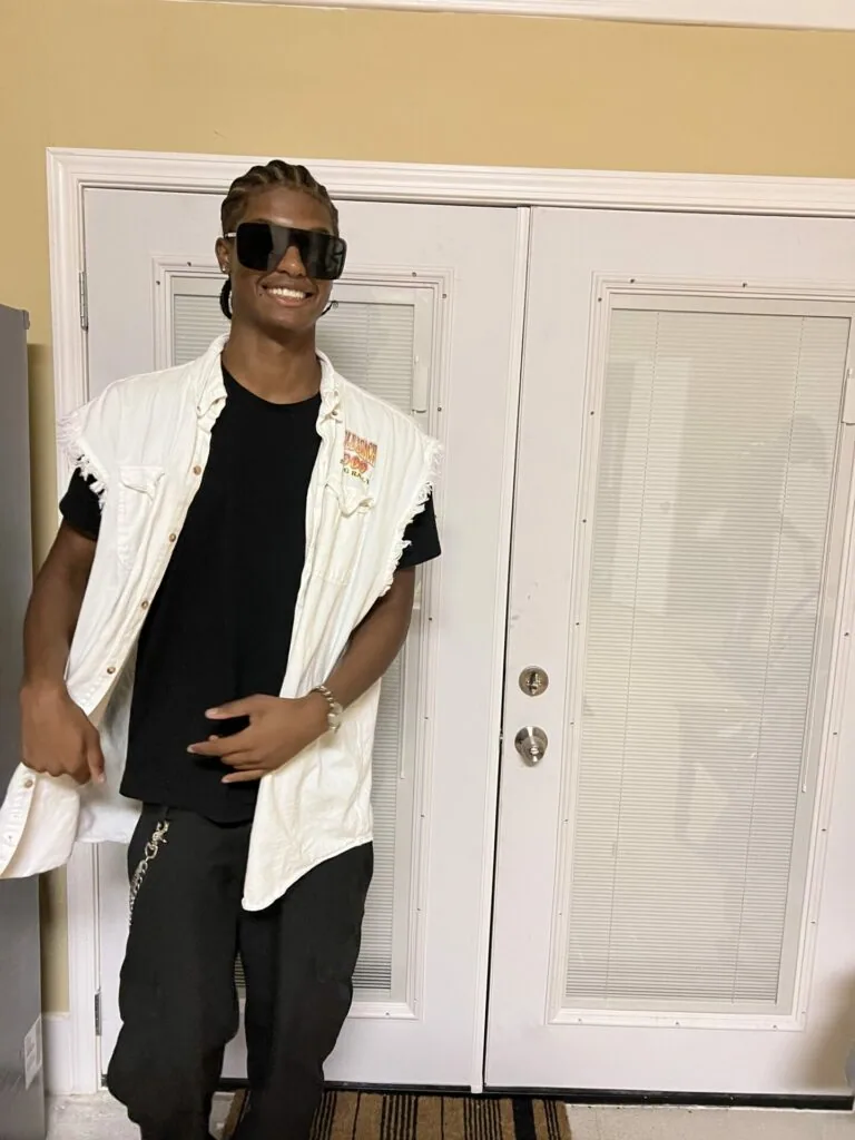 Man wearing white oversized button-up shirt, black t-shirt, sunglasses, posing indoors against white doors