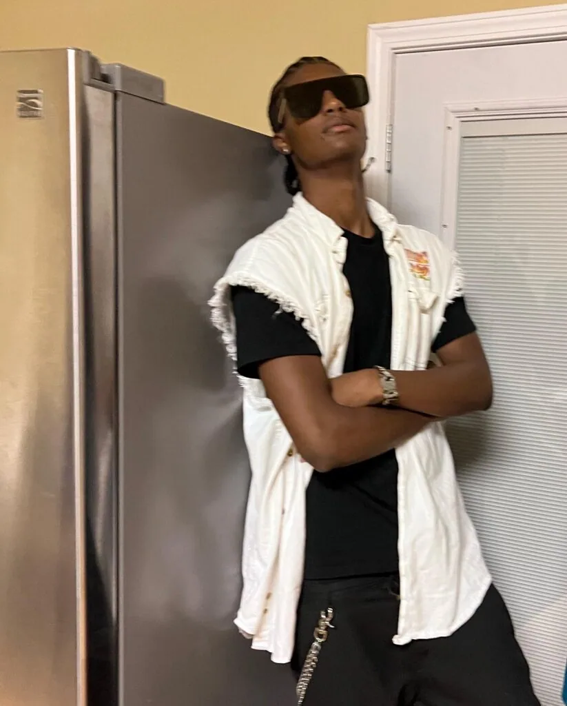 Man in white sleeveless shirt and black tee with sunglasses posing in kitchen by refrigerator