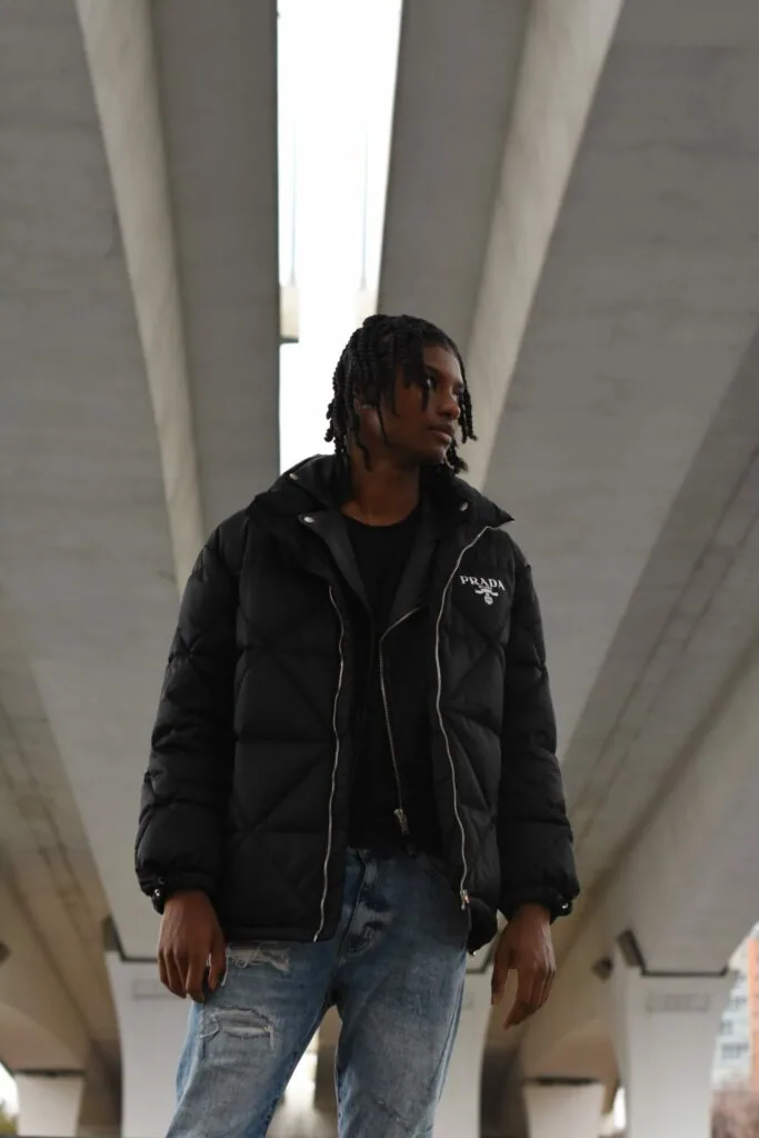 Male model wearing black puffer jacket and distressed jeans in minimalist concrete architectural setting with natural light overhead.