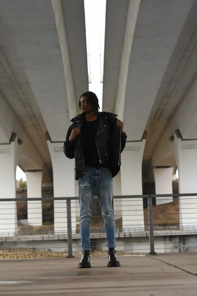 Male model in black leather jacket and distressed jeans standing under concrete archway with natural skylight