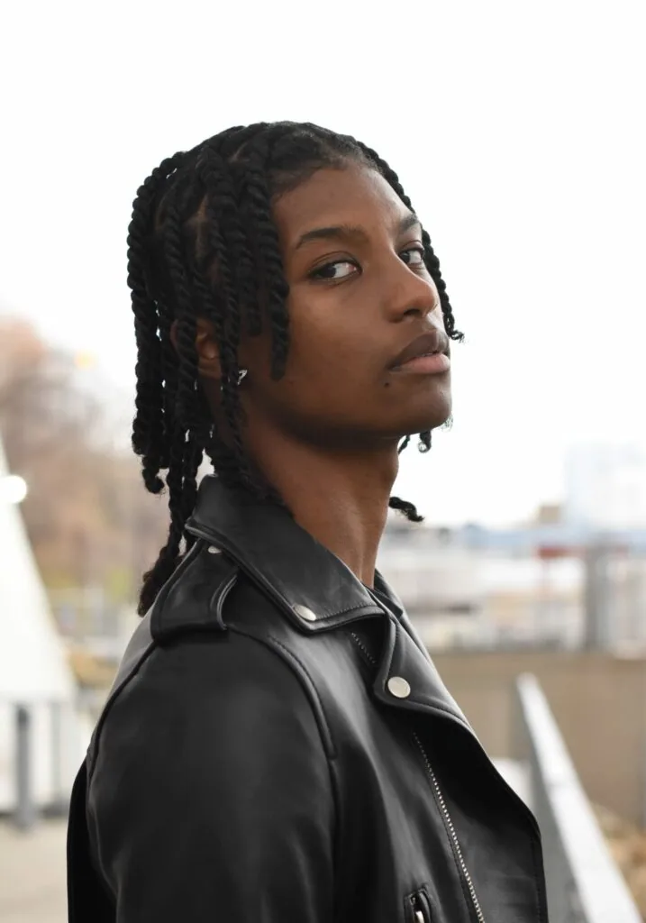 Male model with braided hair wearing black leather jacket on rooftop with city skyline background
