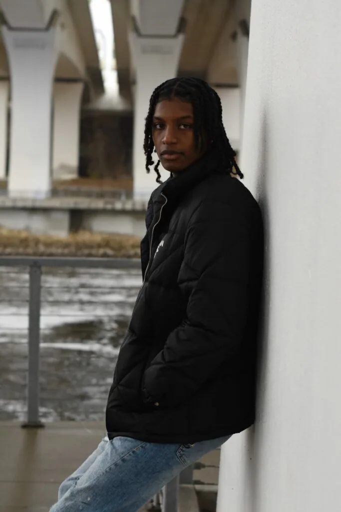 Male model in black puffer jacket and jeans poses against white pillar under modern bridge structure
