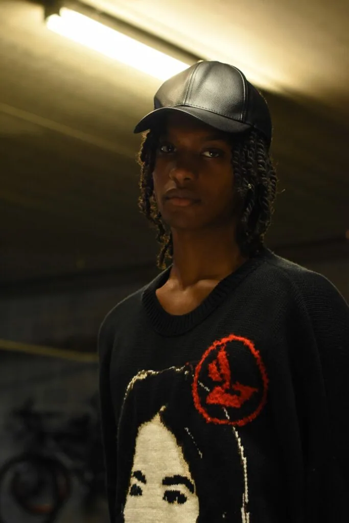 Male model wearing black graphic sweater and gray leather cap in underground garage setting with fluorescent lighting.