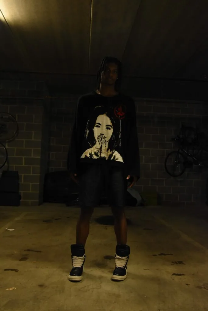 Male model wearing black graphic sweatshirt and high-top sneakers standing in industrial garage setting with brick wall and bicycles