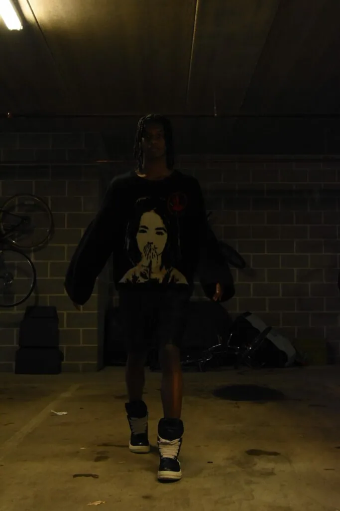 Male model wearing black graphic t-shirt and high-top sneakers standing in industrial brick garage with bicycle and fluorescent lighting