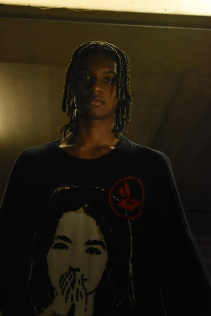 Male model wearing black graphic t-shirt with portrait artwork and red balloon motif, long braids, studio indoor lighting
