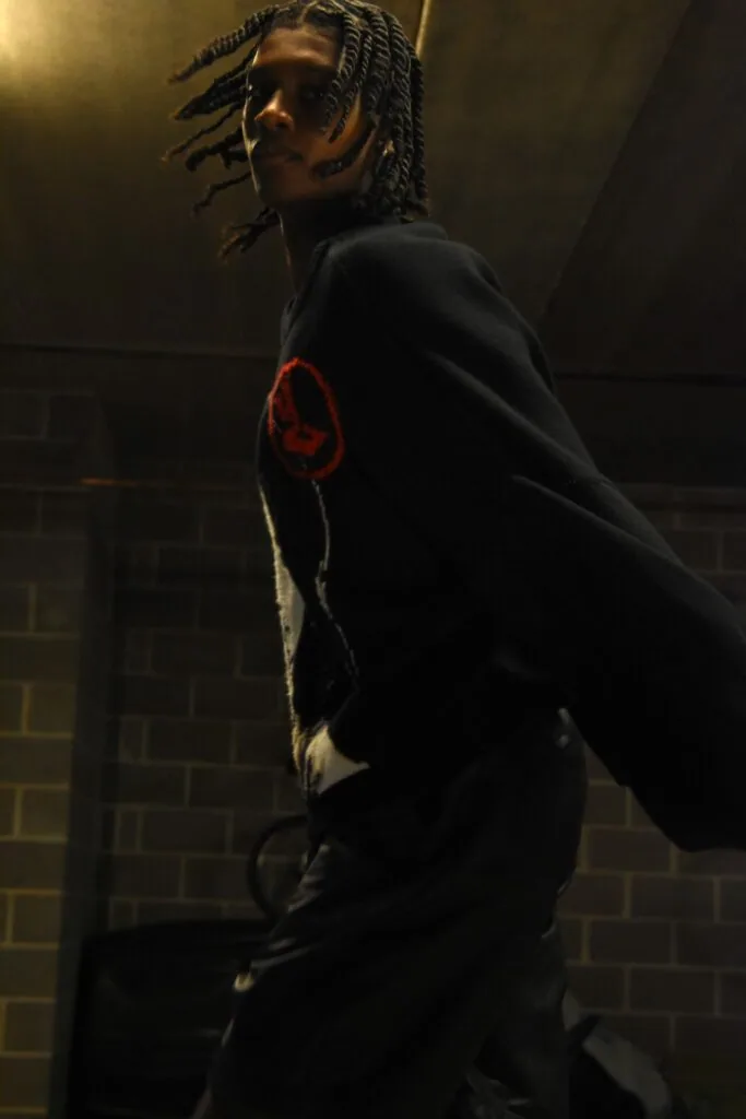 Male model with braided hair wearing black hoodie with red graphic design against brick wall