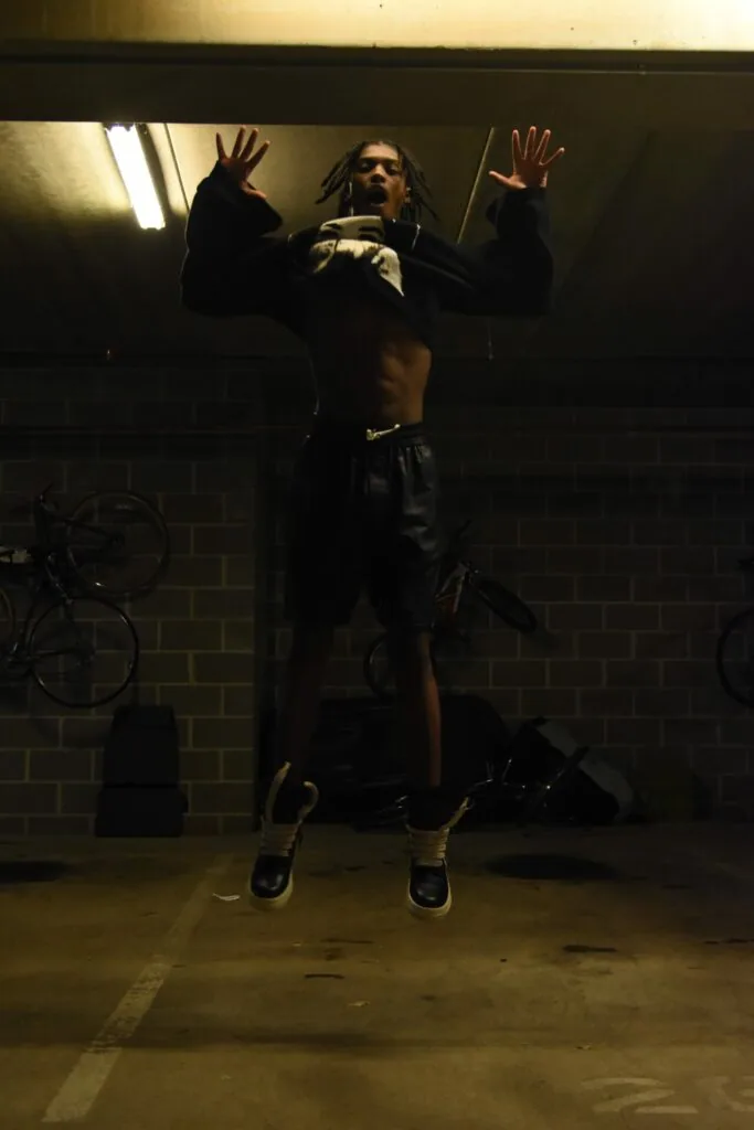 Male model with dreadlocks jumping in black crop top and pants in industrial brick garage with bicycles