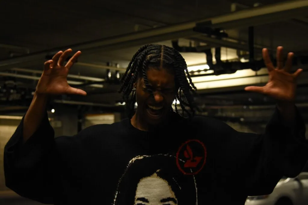 Male model with braids wearing black graphic t-shirt, hands raised, posing in industrial gallery space with warm overhead lighting.