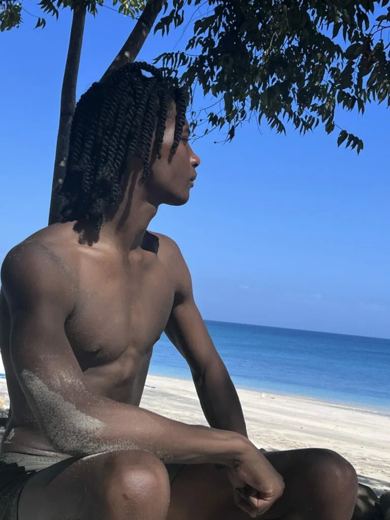 Shirtless male model with braided hair sitting under palm tree overlooking ocean beach