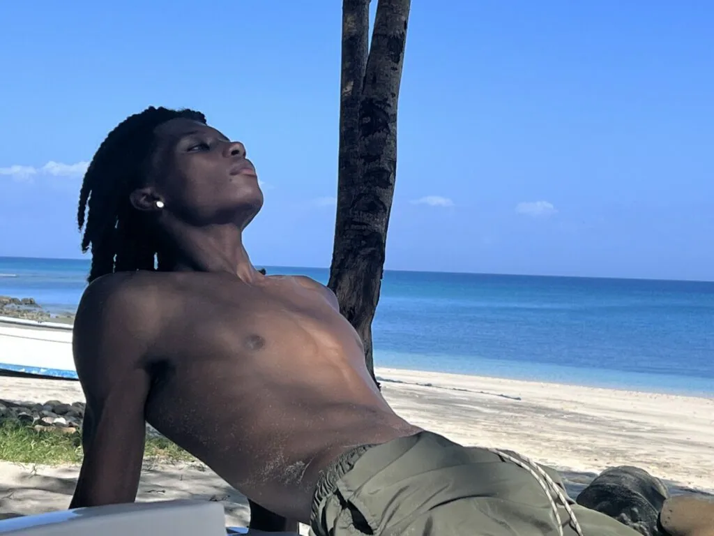 Shirtless man with dreadlocks leaning against palm tree on beach overlooking ocean