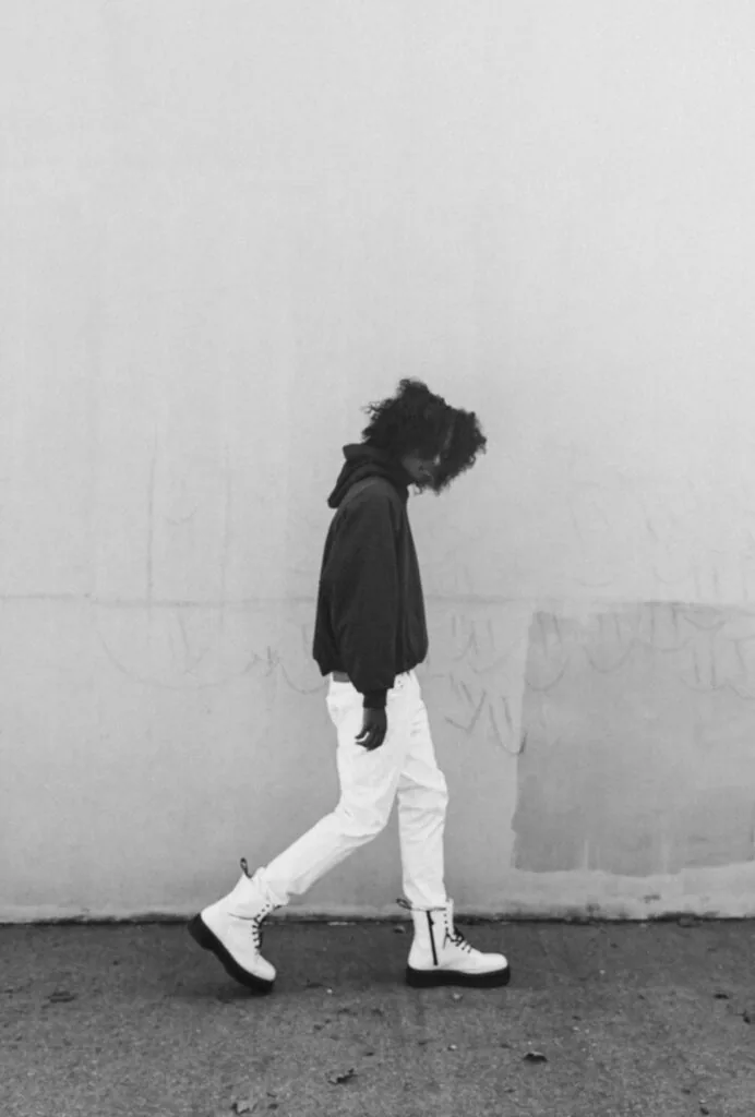 Person with curly hair wearing black jacket and white pants with white sneakers against gray wall