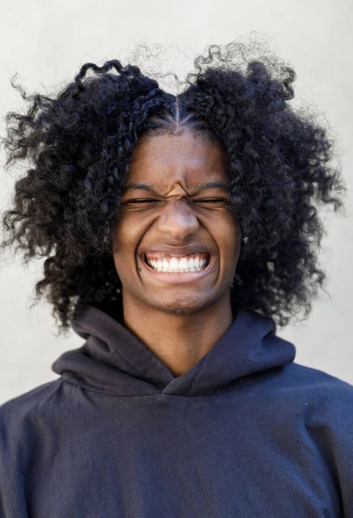 Young man with curly black hair wearing navy hoodie, smiling broadly against white background