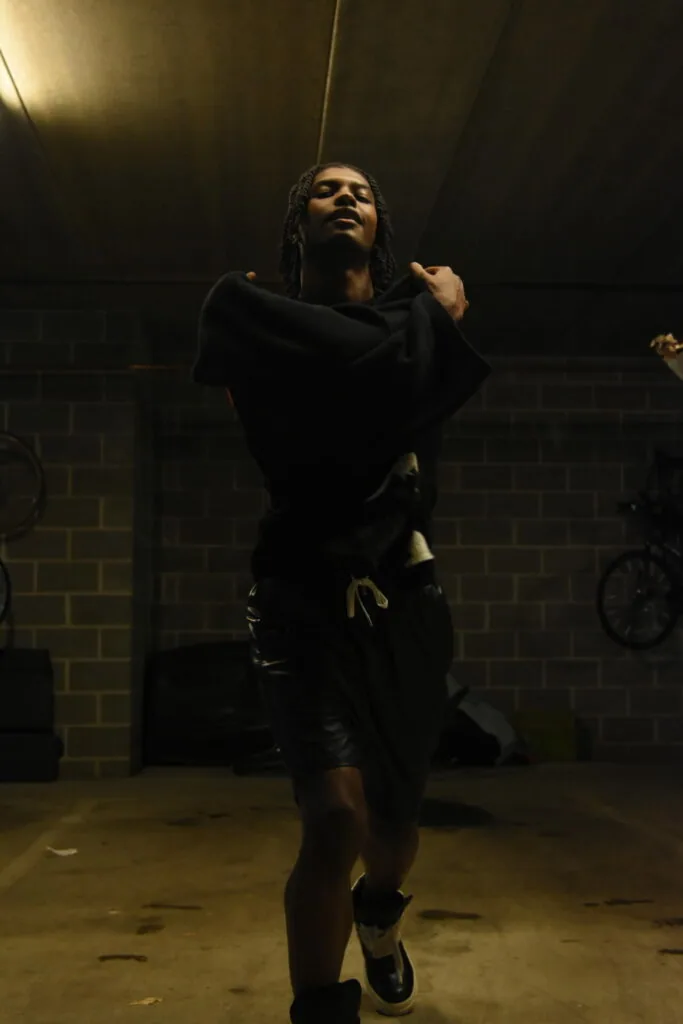 Male model in black hoodie and leather pants posing in industrial brick warehouse setting under warm overhead lighting