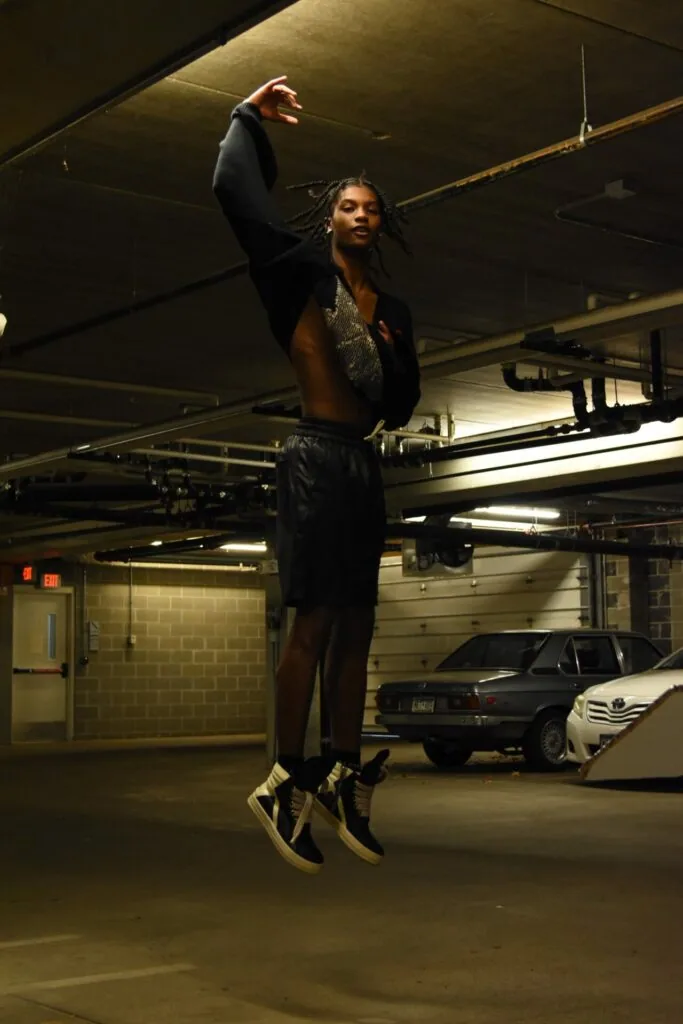 Male model jumping in underground parking garage wearing black blazer and metallic accents with raised fist pose