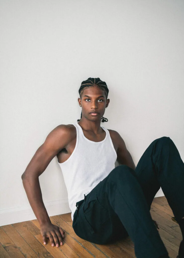 Male model in white tank top and black pants sitting on wooden floor against white wall