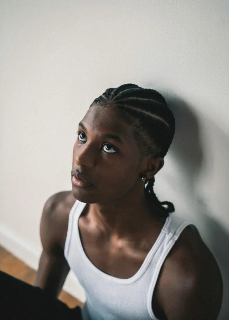 Man with braided hair wearing white tank top against white wall, looking upward thoughtfully
