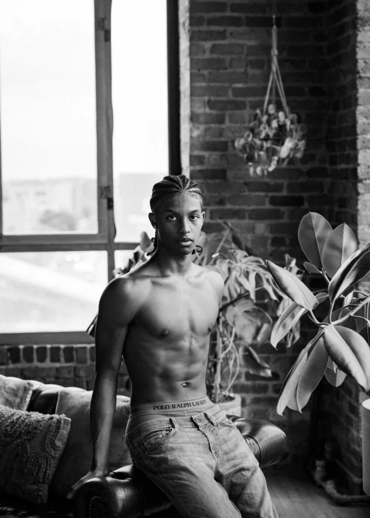 Shirtless male model in jeans sitting by brick window in industrial loft with hanging plants and potted greenery